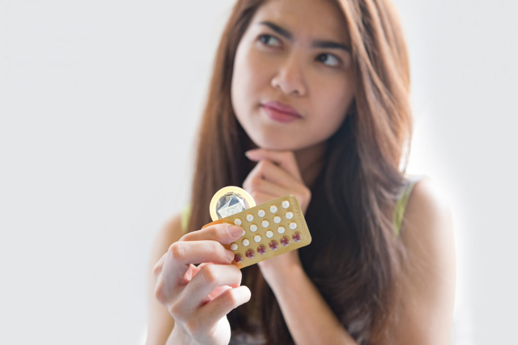 young woman holding a condom and birth control pills while thinking