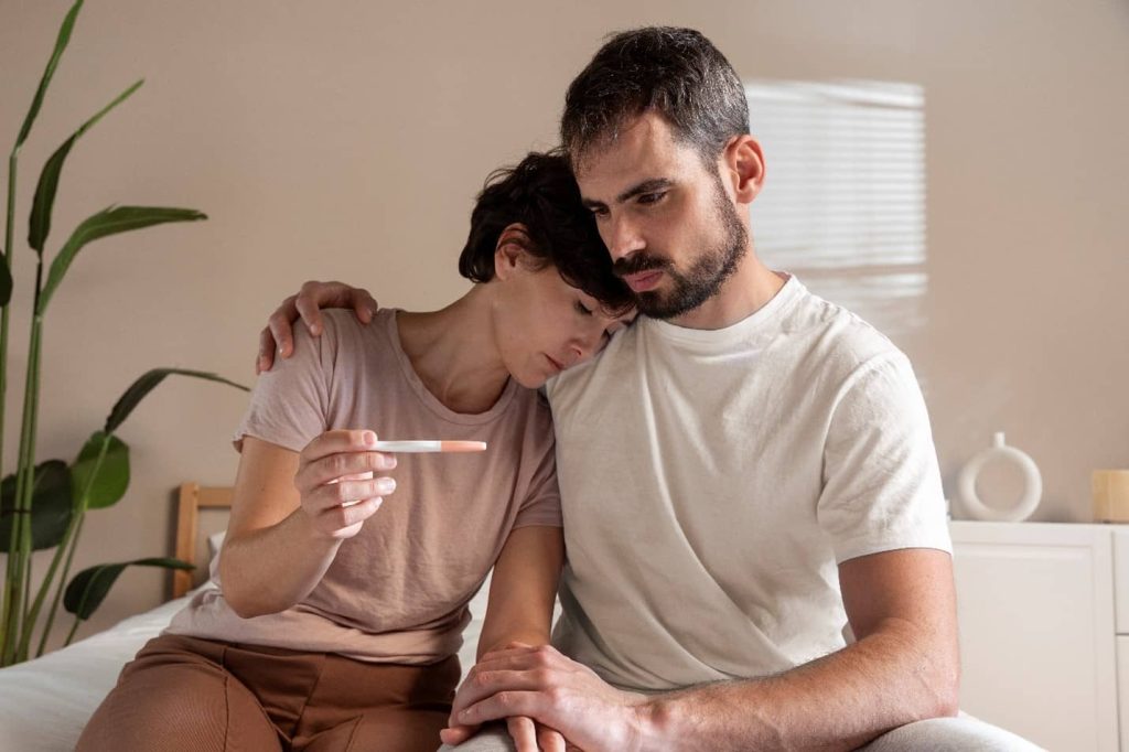 a man and woman sitting on a bed holding a pregnancy test, Can Severe Period Pain Cause Infertility?